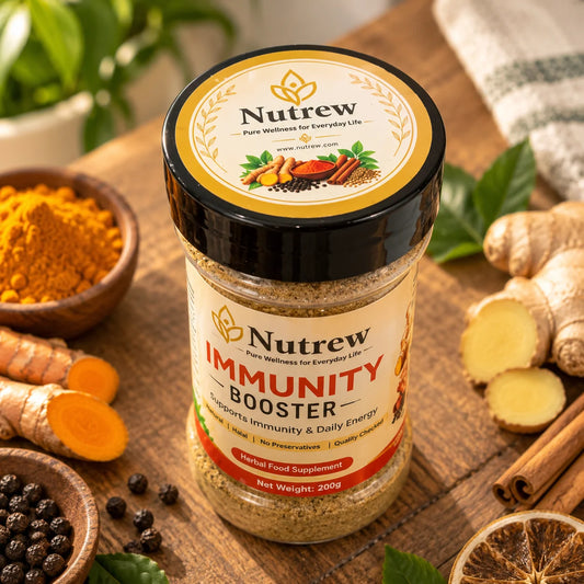 A jar of Nutrew Immunity Booster herbal food supplement placed on a rustic wooden surface, surrounded by ingredients like turmeric root, turmeric powder, ginger, cinnamon, black peppercorns, and dried lemon slices. The background is a warm, softly blurred kitchen setting with green leaves and a striped cloth.