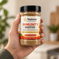 A hand holding a clear plastic jar of Nutrew Immunity Booster herbal food supplement, with a warm, softly blurred indoor background.