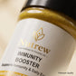 Close-up of a Nutrew Immunity Booster jar showing the white label with gold and black text, a gold powder visible at the top, and a black lid against a soft beige background.