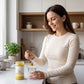 A woman in a cream ribbed long-sleeve top stands in a bright kitchen, holding a spoon with a yellow powder over a glass of water next to a jar of 'Yellow Turmeric' capsules. The jar label features a gold logo and text.