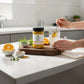 Person using a jar of honey on a kitchen counter with oranges and herbs.